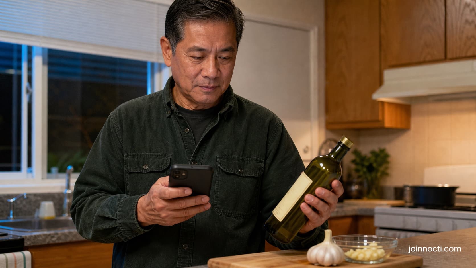 Adult man in a home kitchen comparing a plain bottle of garlic-infused olive oil with a recall notice on his phone