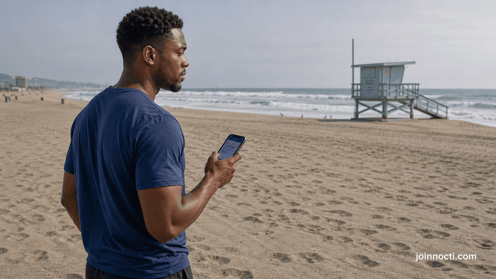 Man checking a weather alert from dry sand while looking toward breaking waves and a lifeguard tower at a Los Angeles County beach