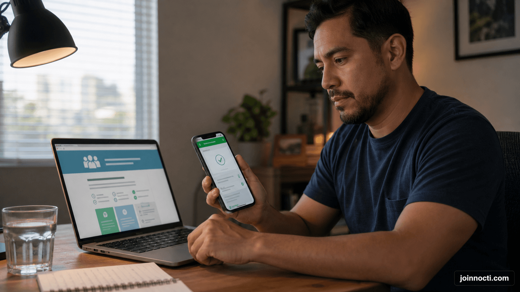 Latino man at a home desk comparing charity verification information on a laptop and smartphone before donating