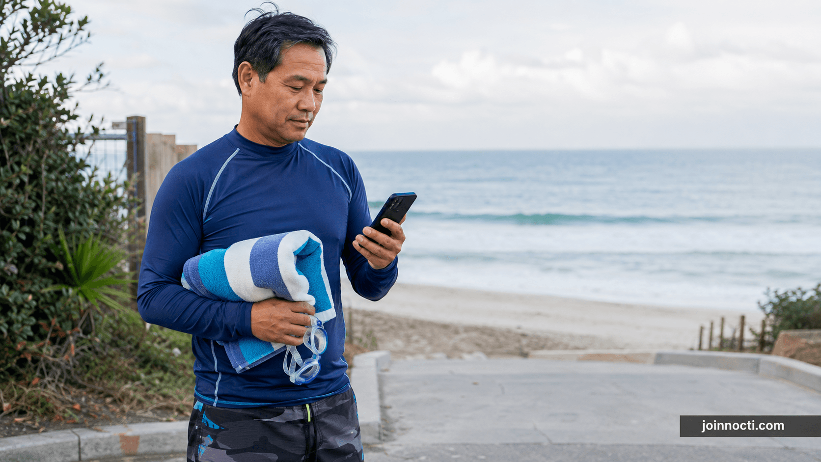 Man in a long-sleeve rash guard checking a beach water-quality update before entering the ocean