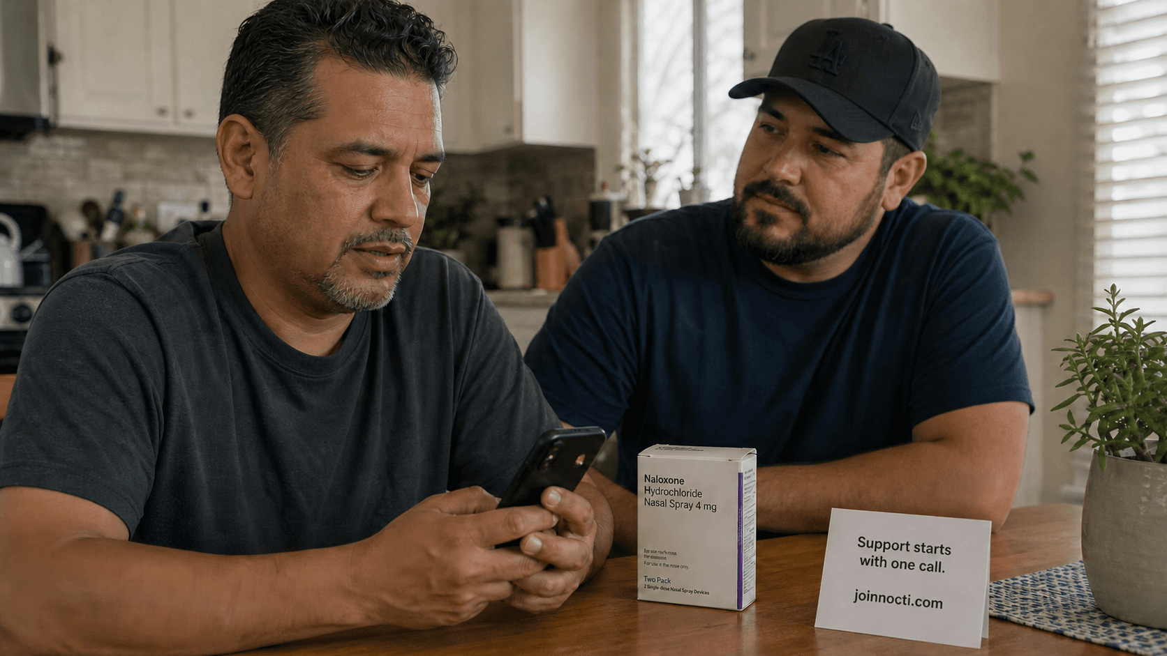 Two Latino men at a kitchen table reviewing an opioid-treatment support resource beside a generic naloxone nasal-spray carton