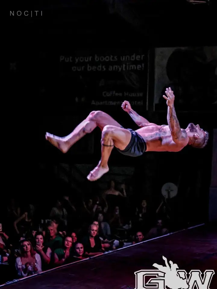 Adult male performer in motion above a stage, illustrating the work and risk behind independent professional practice; original event mark remains visible